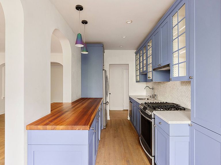 Galley kitchen of 360 Clinton 3T with periwinkle blue custom cabinetry, butcher block island, gas range, and pendant lighting under an arched opening